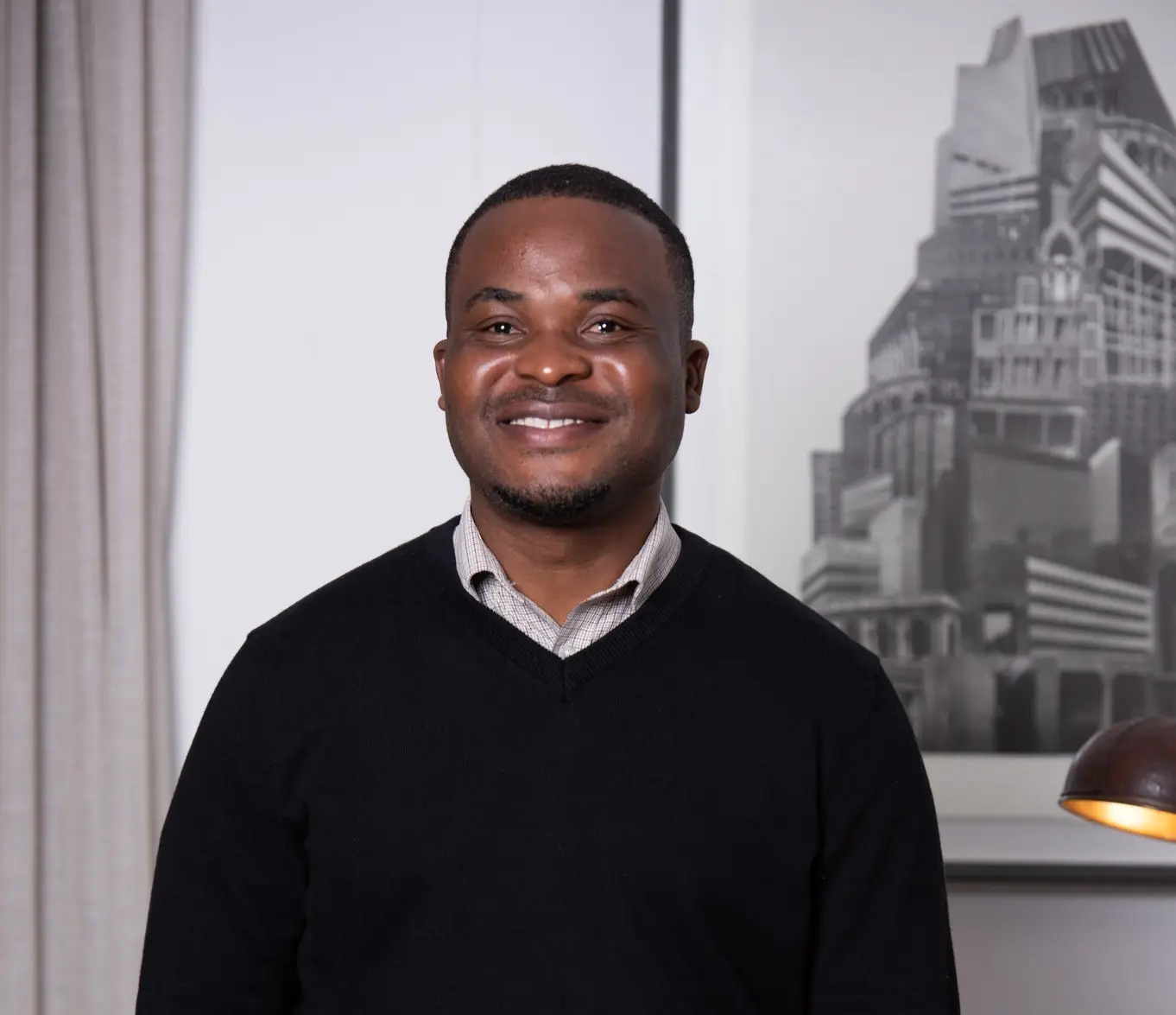 Portrait of a smiling man wearing a black V-neck sweater over a light checkered shirt, with a grayscale cityscape artwork in the background.